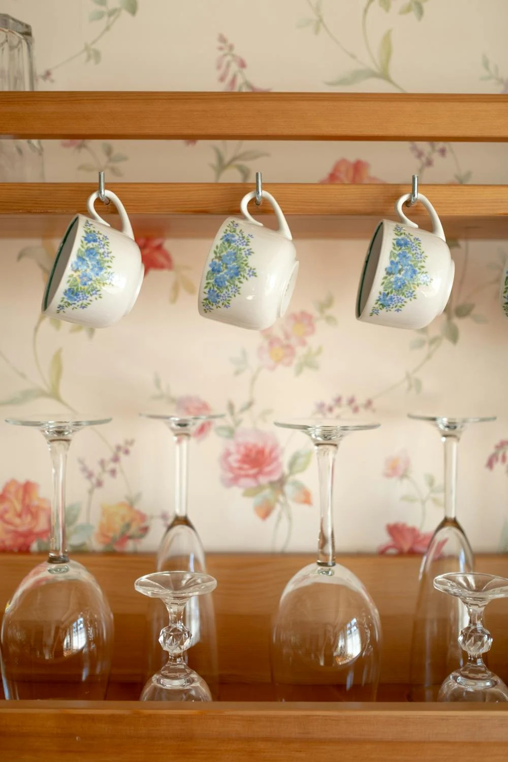 An old fashioned kitchen with floral wallpaper and hanging floral teacups. There are clear, upside-down glasses as well.